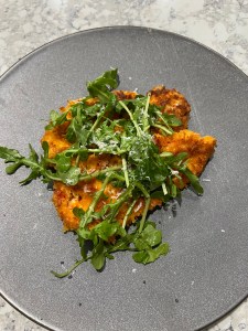 A plate with a crispy fritter topped with fresh arugula and grated cheese, set against a dark gray background.